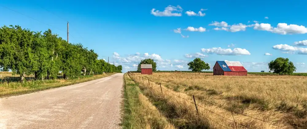An abandoned old barn in Rural Texas with the state flag painted on its roof sits in a farmland community framed by fields and a dirt road. An abandoned old barn in Rural Texas with the state flag painted on its roof sits in a farmland community framed by fields and a dirt road.